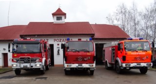 trzy samochody pożarnicze, remiza strażacka w tle